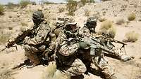 Vier bewaffnete Soldaten hocken im Kreis mit dem Rücken zueinander in der Wüste