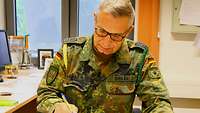Ein Soldat sitzt am Schreibtisch in seinem Büro und schreibt mit einem Kugelschreiber auf ein Blatt Papier.