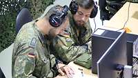Zwei Soldaten mit Kopfhörern sitzen nebeneinander an einem Tisch, einer schreibt, vor ihnen Rechner und Bildschirm.