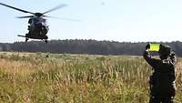 Ein Soldat steht auf einer Wiese und hält eine signalgrüne Tafel hoch. Vor ihm landet gleich ein Hubschrauber.