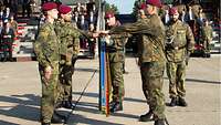 Soldaten stehen links und rechts an einer Truppenfahne und halten die linke Hand darüber.