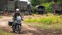 Ein Soldat auf einem Motorrad fährt durch das Gelände, dahinter stehen mehrere Lkw. .