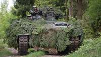 Ein mit Zweigen getarnter Panzer steht im Wald. Aus der Luke des Panzers schaut ein Soldat.