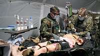 Zwei Soldaten stehen an einem Feldoperationstisch in einem Sanitätszelt und versorgen eine liegende Übungspuppe 