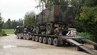 Ein Lastkraftwagen steht auf einem großen Schwerlasttransporter und wird mit Ketten von einem Soldaten gesichert.
