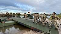 Soldaten marschieren über ein grünes, metallenes Brückenteil, das am Flussufer aufliegt.