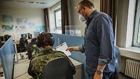 Ein Mann steht neben einem Soldaten, der vor einem Computer sitzt, und zeigt ihm einen Zettel