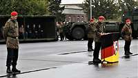 Mehrere Soldaten in Uniform stehen draußen auf einem Platz inmitten von Gästen bei einem Appell. 