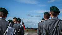 Eine Formation von Soldaten steht mit Blickrichtung zum Hintergrund des Bildes. Ihnen kommen am Himmel vier Flugzeuge entgegen.