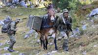 Zwei Soldaten bringen mit einem bepacktem Maultier Ausrüstung über eine felsige Bergwiese den Berg rauf.