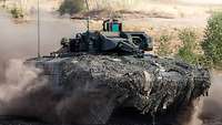 A camouflaged armoured fighting vehicle is driving fast; dust is being whirled up.