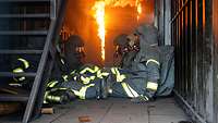 Mehrere Personen in voller Brandschutzausrüstung sitzen in einem Container und beobachten züngelnde Flammen im Hintergrund.