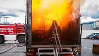 Person in voller Brandschutzausrüstung hockt in einem Container, hinter ihr baut sich eine Flammenwand auf.