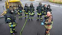 Männliche Person in Arbeistkleidung gibt Erläuterungen an mehrere Personen, die volle Brandschutzausrüstung tragen.