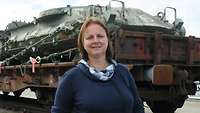 Frau in blauer Kleidung steht vor einem Eisenbahnwaggon mit Panzern