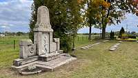 Ein mannshoher Grabstein neben mehreren liegenden Grabsteinen auf einer Wiese