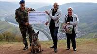 Drei Personen und ein Hund stehen vor Bergkulisse. Ein Soldat hält mit einem Mann den Spendencheck hoch.