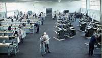 A great number of men are working at workbenches in a large hall with a stone floor