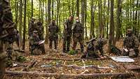 Mehrere Soldaten stehen im Wald vor einem Geländesandkasten, gelegt aus Ästen und farbigem Band.