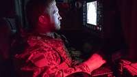 Ein Soldat sitzt in einem geschützten Fahrzeug in Schutzweste vor einem Monitor und bedient einen Joystick