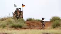 Auf einer roten Sandpiste steht ein Dingo mit der deutschen Flagge sowie der Flagge der Vereinten Nationen, rechts ein Soldat 