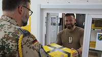 Ein Soldat gibt an einem Schalter ein Postpaket auf. Der Feldpostsoldat nimmt dieses entgegen.