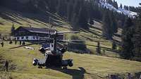 Eine CH-53 steht mit geöffneter Heckklappe auf einer Alm. Im Hintergrund sieht man Mitglieder des THW und eine Alphütte.