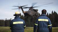 Zwei Kameraden des THW tragen Uniformen mit der Rückenaufschrift „THW“ und schauen einer CH-53 beim Landen zu.