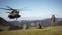 Drei Soldaten knien auf einer Wiese und sehen zu einem Hubschrauber, der sich im Anflug befindet.