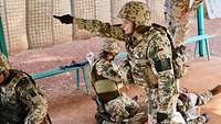 Eine Soldatin zeigt mit dem Finger in eine Richtung, um sie herum liegen sternförmig die Verletzten, Ersthelfer knien davor