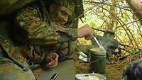 Ein Soldat liegt unter einer aufgespannten Zeltplane und kocht sich im Feldgeschirr etwas.