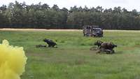 Auf einem Feld steigt gelber Rauch auf, drei Soldaten ducken sich. Im Hintergrund fährt ein militärisches Fahrzeug.