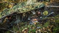 Soldaten sitzen unter einer Zeltplane im freien Gelände und kochen an einer offenen Feuerstelle