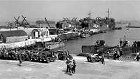 Schwarzweißaufnahme eines Hafens mit Schiffen, Landungsbooten und einer Reihe von Jeeps auf einer Pier.