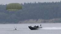 Ein Soldat landet mit einem Fallschirm im Wasser. Rechts von dem Soldaten fährt ein Schlauchboot mit Soldaten an Bord.
