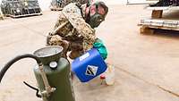 Im Vordergrund eine olivfarbene Kübelspritze, dahinter kniet ein Soldat mit Maske und füllt Flüssigkeit in einen Messbecher ab