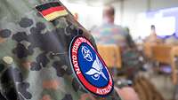 Das rot-blaue Patch mit weißer Schrift, einem stilisierten Eulenkopf und dem NATO-Logo am Uniformärmel eines deutschen Soldaten
