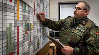 A soldier with a writing pad is standing in an office at the planning chart.