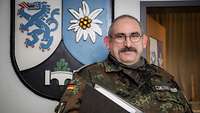 A soldier with a folder under his arm stands in front of a big coat of arms on the wall.