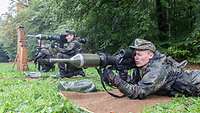 Two soldiers, kneeling and lying respectively, hold shoulder-fired antitank weapons at the ready near the edge of a wooded area.