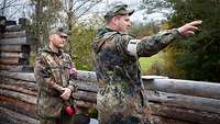 Zwei Soldaten stehen hinter einer halbhohen Wand aus Holzbalken. Einer weist mit dem Arm ins Gelände