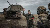 Eine Soldatin steht mit erhobenem Arm im Gelände vor einer Panzerhaubitze 2000 und erkärt Soldaten etwas