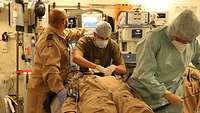 Zwei Mediziner und ein Anästhesiepfleger stehen um eine Trage, auf der ein Mann liegt. Ringsherum stehen medizinische Geräte.