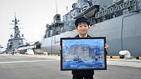 Ein japanischer Junge steht im Hafen vor einem Kriegsschiff und hält ein Bild hoch