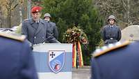 Soldat steht am Rednerpult. Im Hintergrund stehen Ehrenposten neben dem Gedenkstein und einem Kranz.