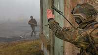 Ein Soldat hinter einem Gebäude gibt einem anderen rennenden Soldaten ein Handzeichen. 