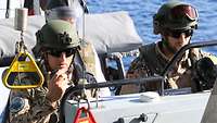 Zwei Soldaten stehen am Steuer auf einem Boot. Ein Soldat hält ein Funkgerät in seiner Hand.