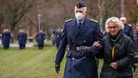 Ein Soldat in blauer Uniform führt eine ältere Dame am Arm.