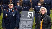 Ein Soldat und eine Frau stehen links und rechts von einer Stele zu Ehren Jürgen Schumanns.