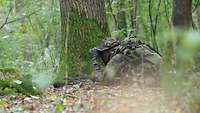 Ein Soldat liegt getarnt mit seiner Waffe im Wald neben einem Baum und schaut durch sein Fernglas. Er hat einen Rucksack auf.